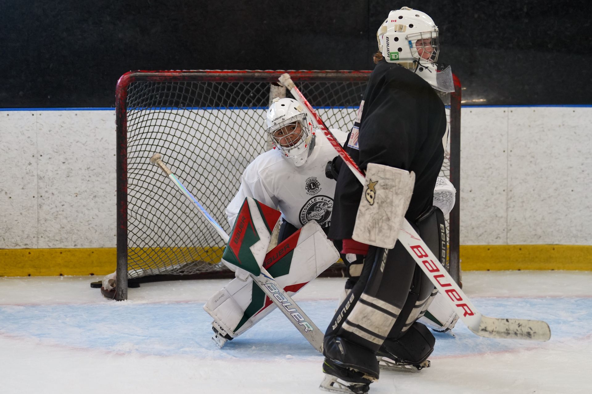Hockey goalie in white blocks a shot as a player in black stands near the net