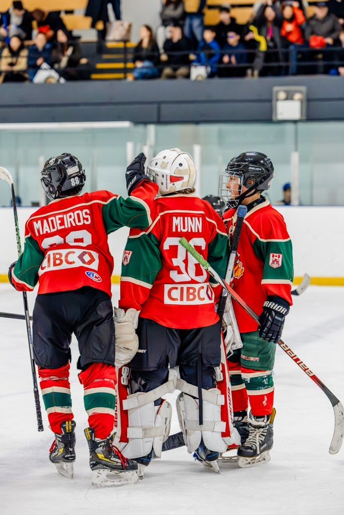 Three hockey players in red and green jerseys huddle on the ice, seen from behind.