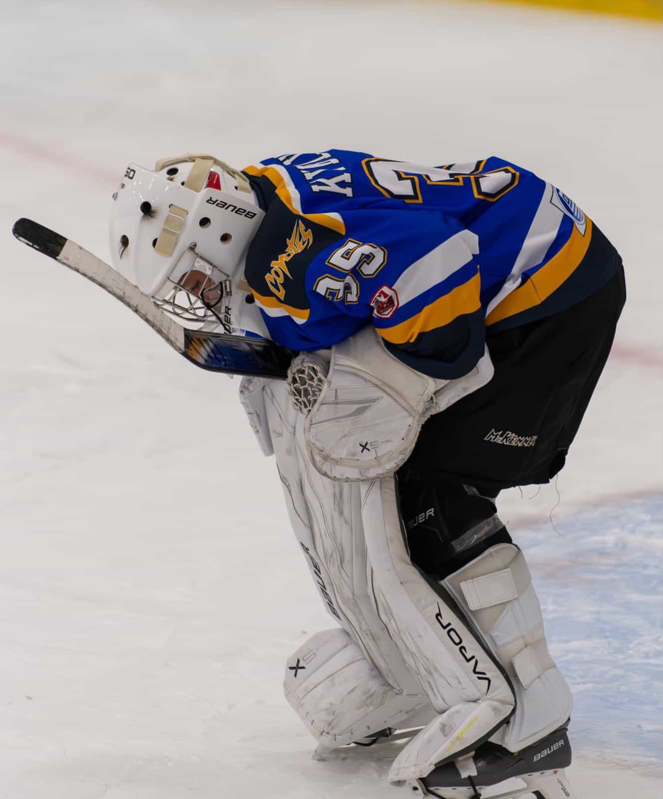 Hockey goalie in white pads and mask crouches while being checked by a blue jersey player