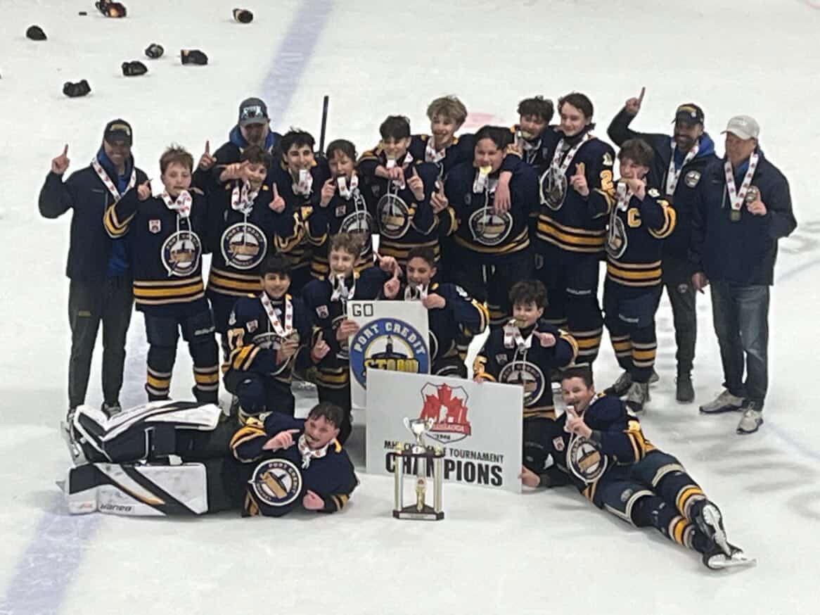 Youth hockey team posing on the ice with championship trophy and banner, wearing dark uniforms.