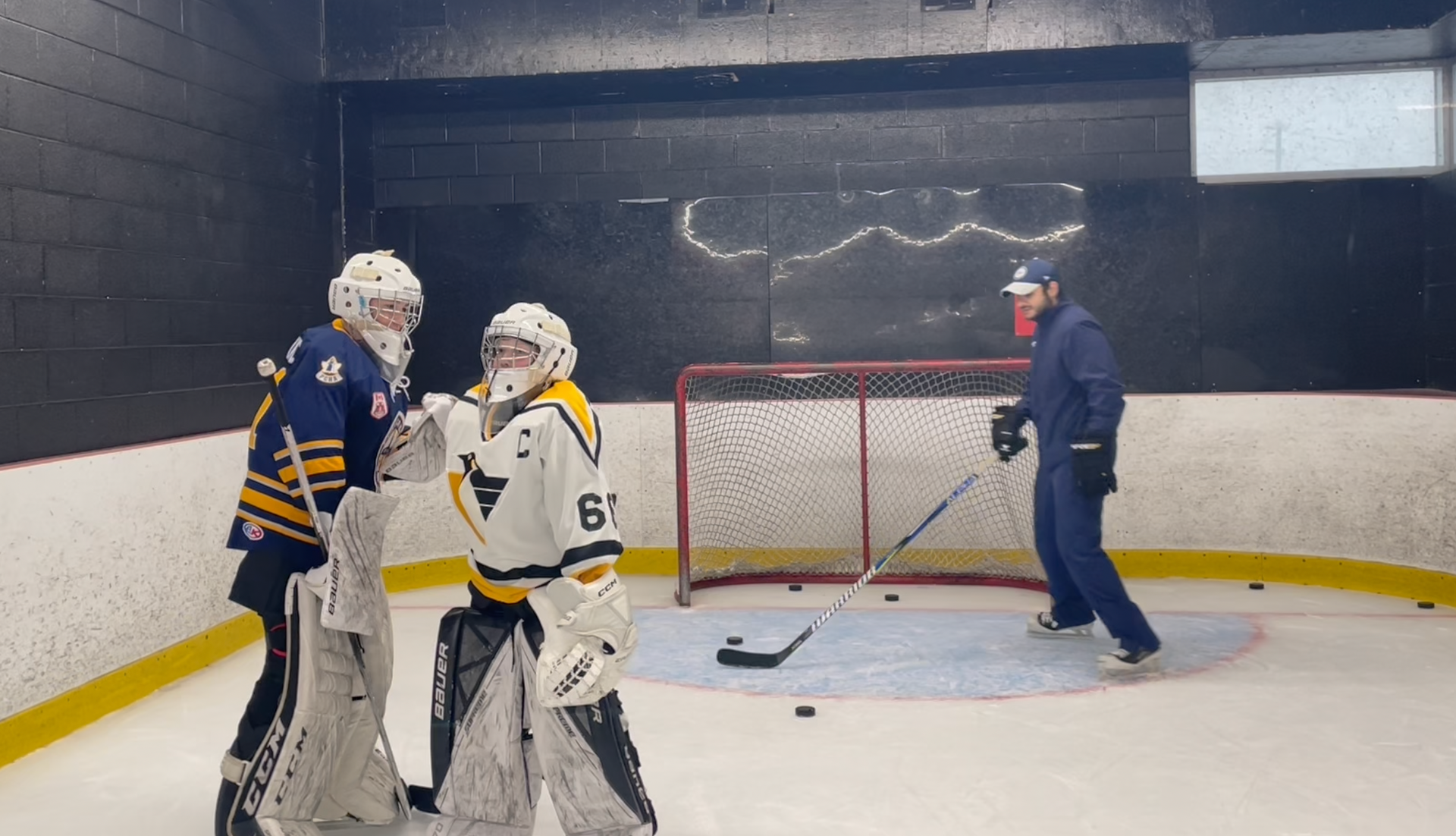 Hockey players and a coach on an indoor rink near a goal, with pucks on the ice.