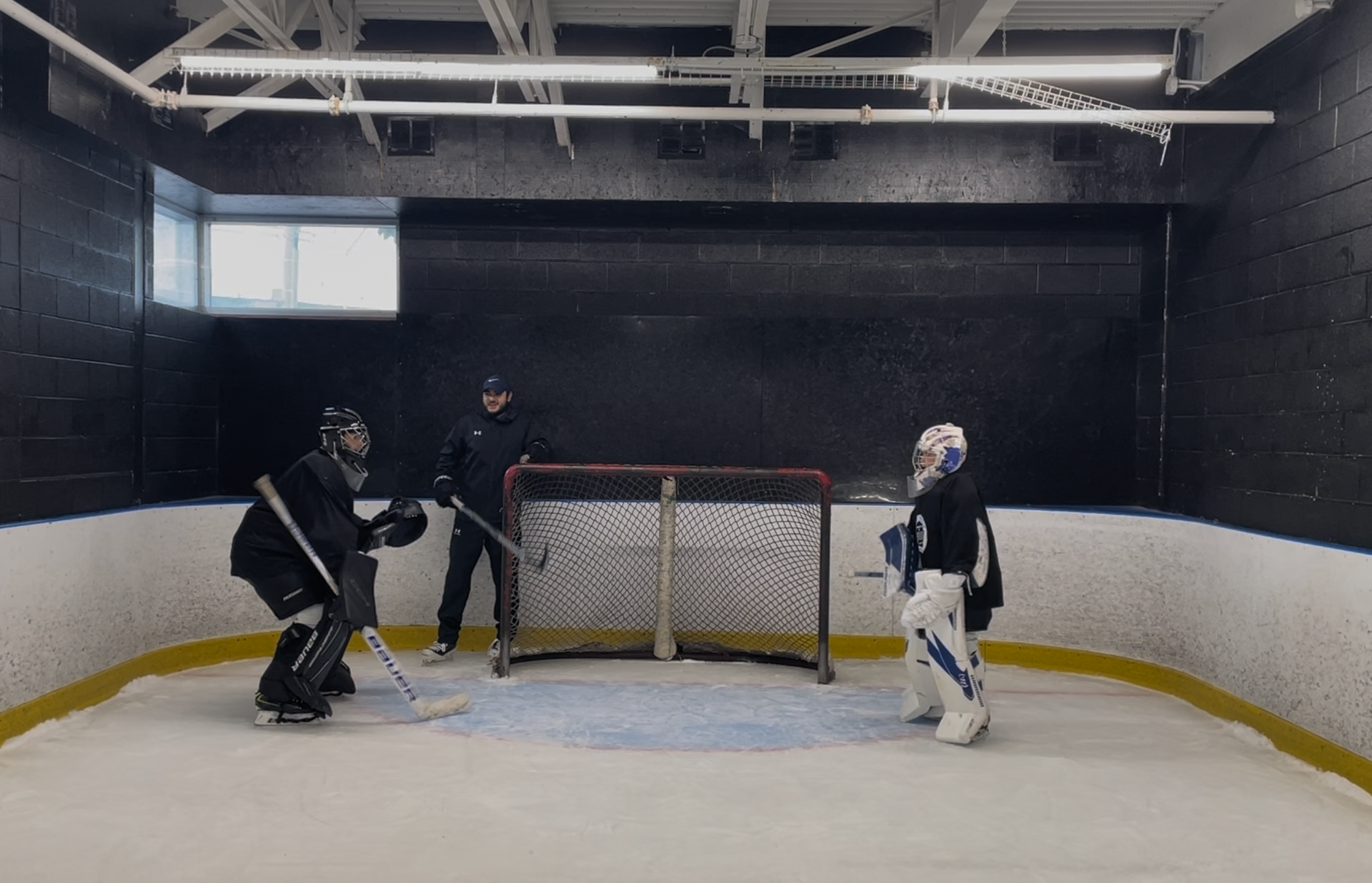 Two hockey players and a goalie on an indoor rink, facing a net in a black-walled practice area