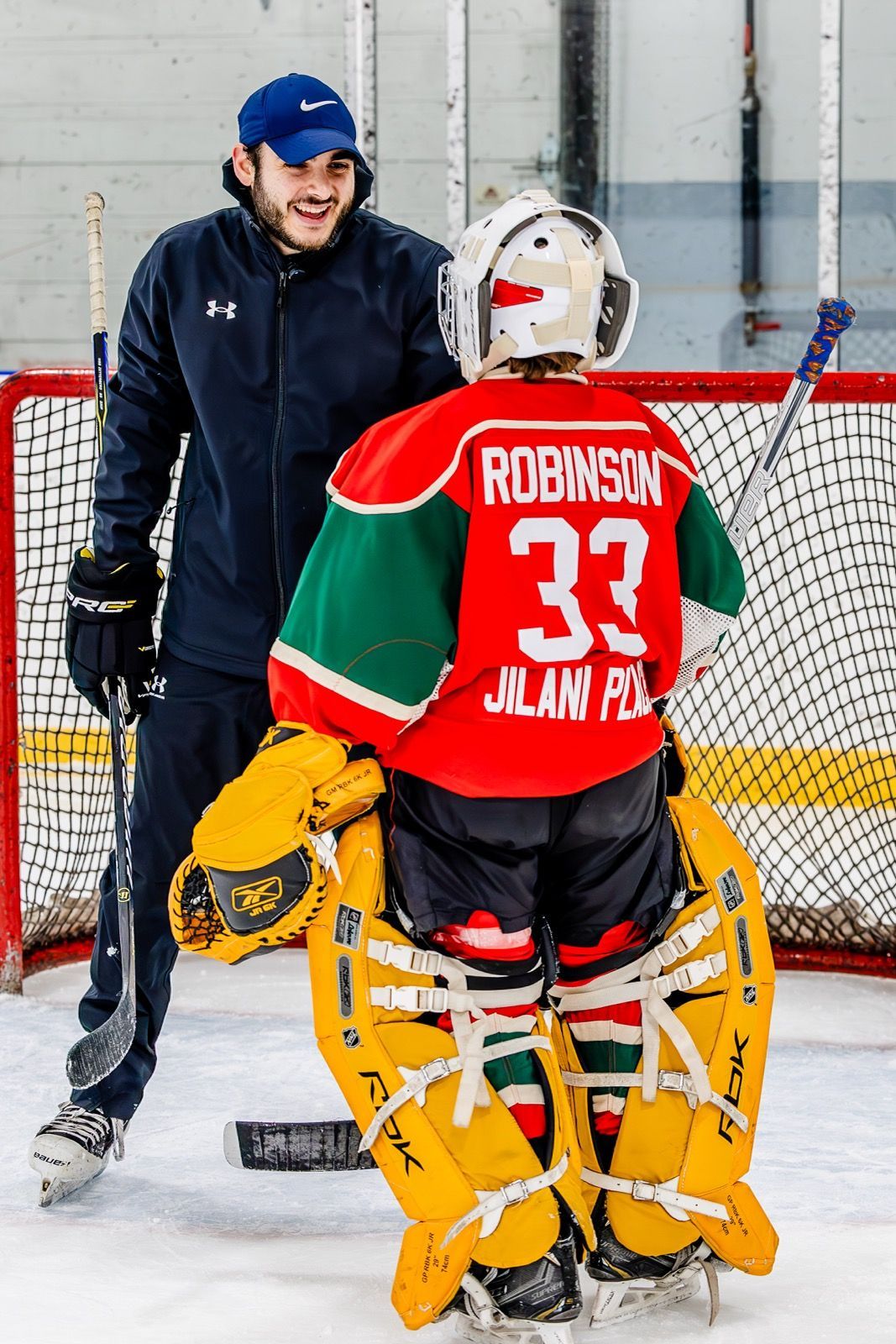 Hockey player in red jersey with goalie gear stands facing coach on indoor rink in front of net.