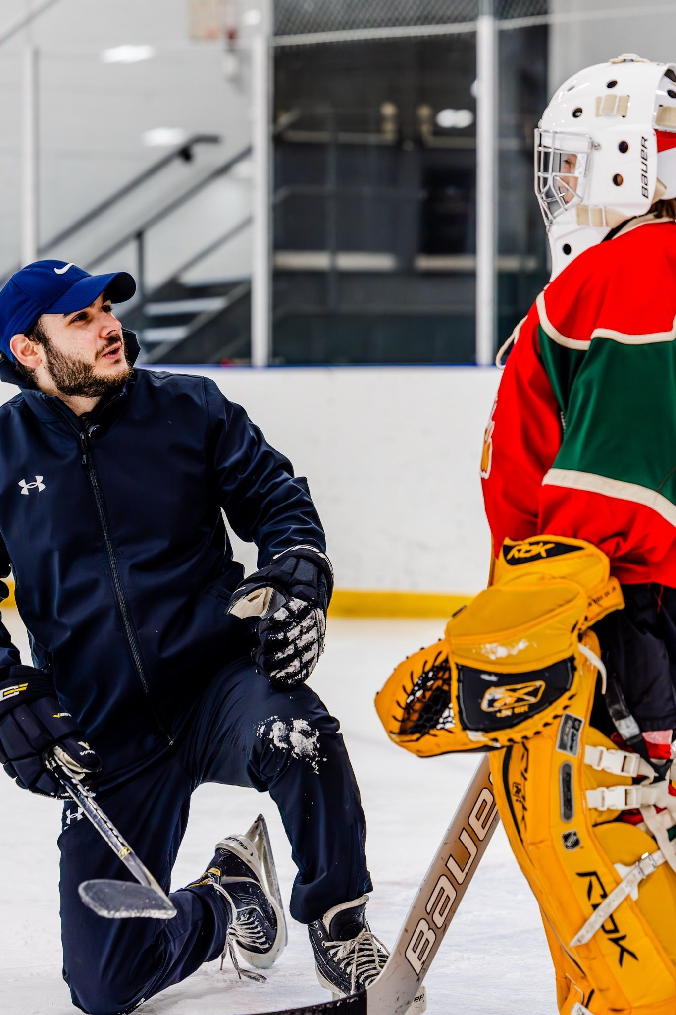 Coach kneels on ice talking to goalie in red and green hockey gear
