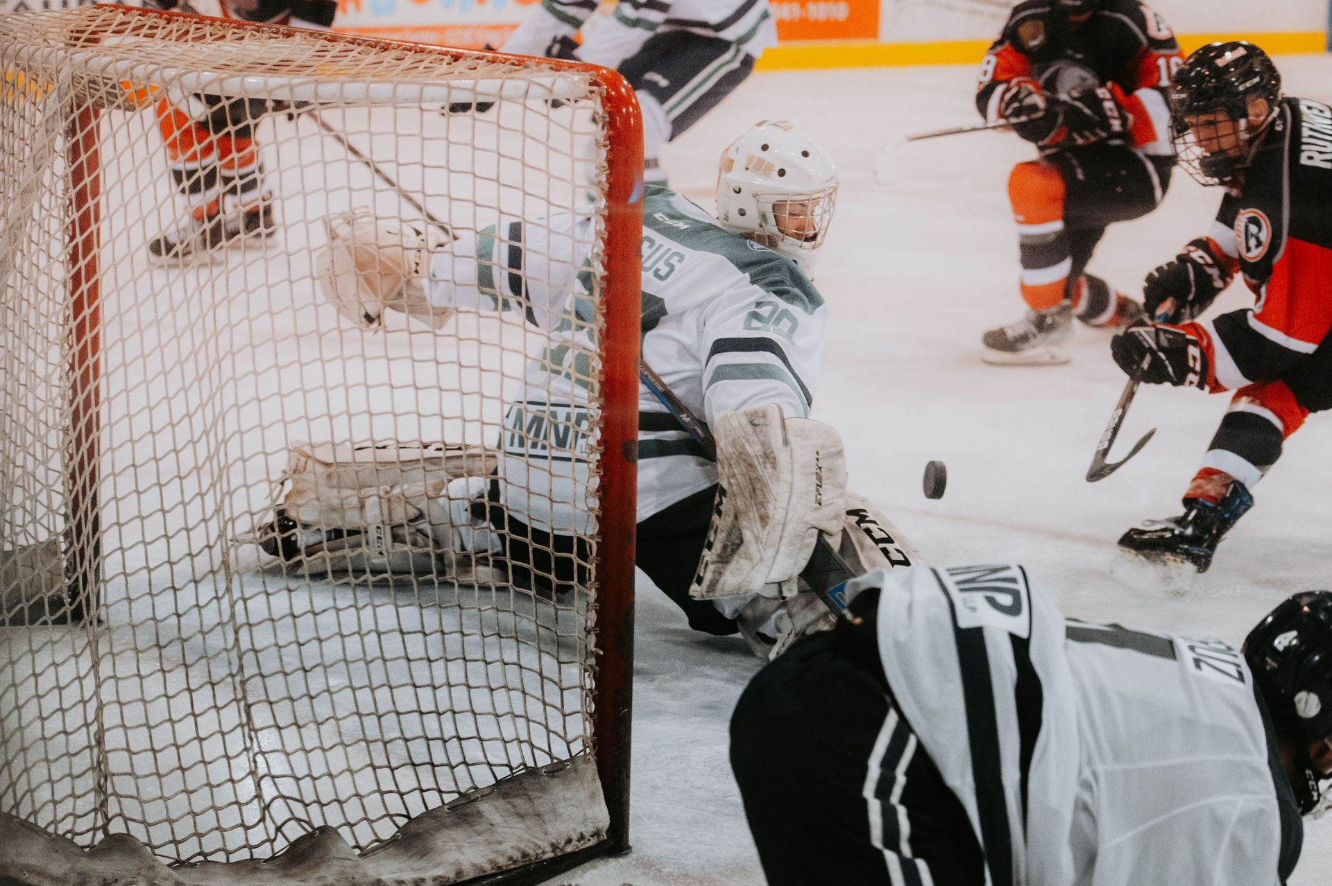 Hockey goalie sprawls to block a puck as players battle near the net on ice rink