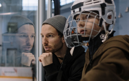 Two men at an indoor ice rink, one in a hockey helmet, both watching the game intently.