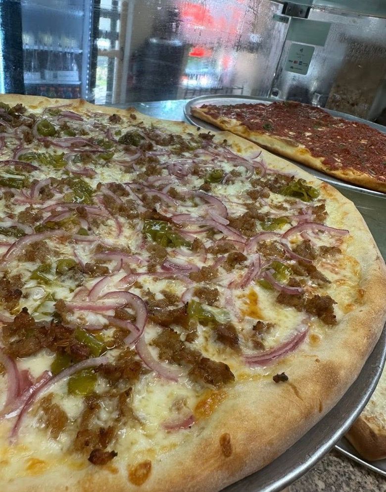 Two pizzas on metal trays, one topped with sausage and onions, another with red sauce in the background