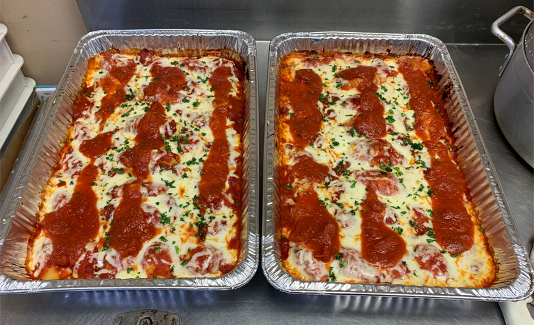 Two foil pans of baked lasagna topped with red sauce, cheese, and herbs on a kitchen counter