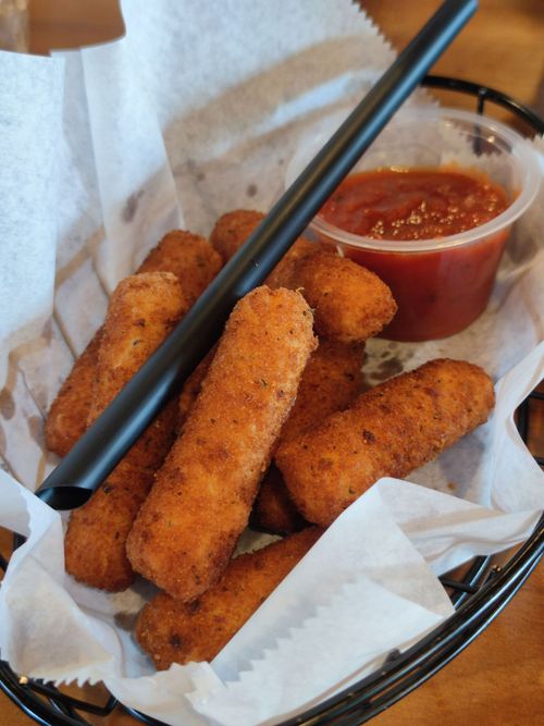 Breaded mozzarella sticks in a basket with marinara dipping sauce and a black pair of tongs