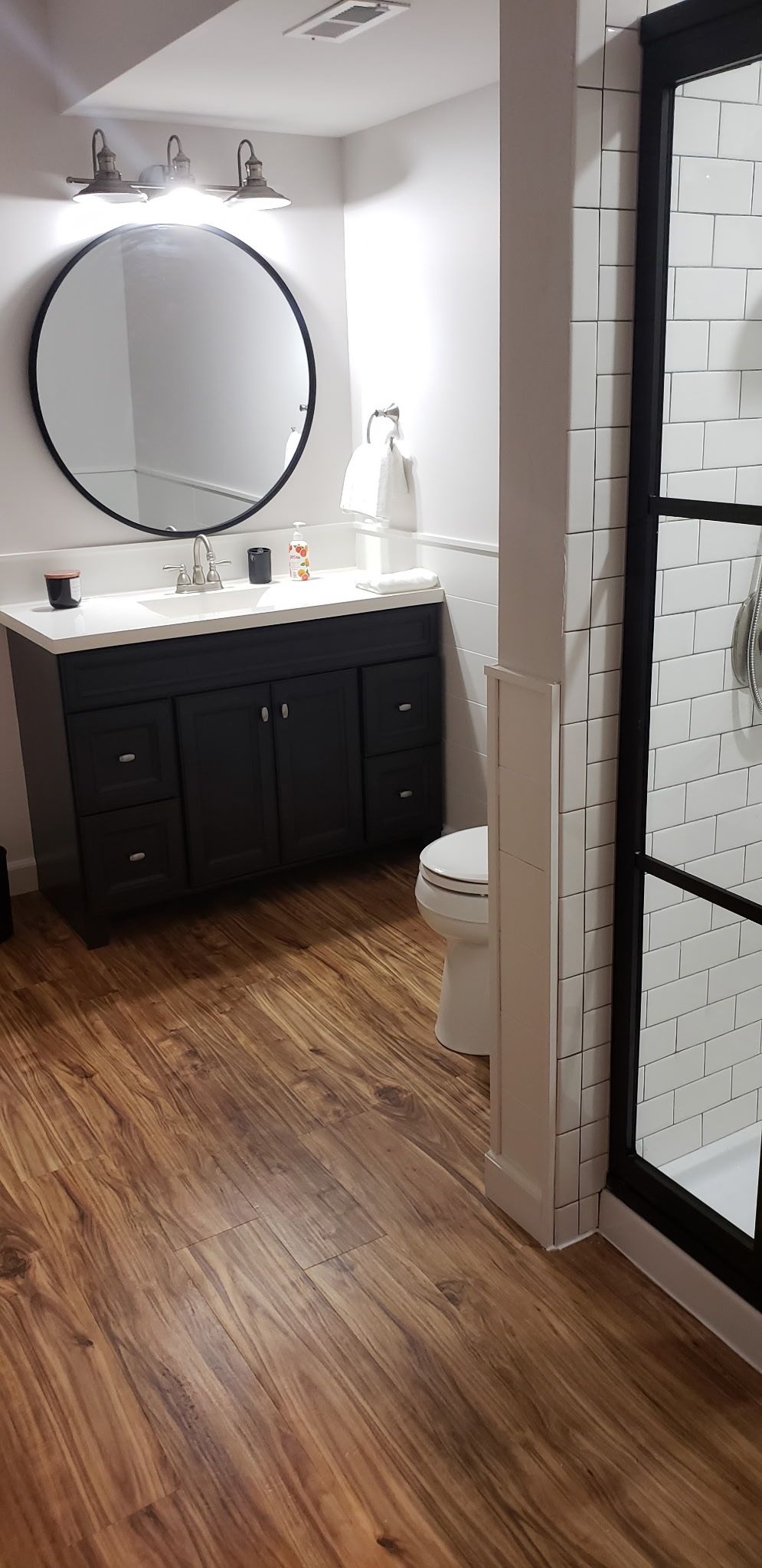 Modern bathroom with round mirror, black vanity, and glass shower door; white tile walls and wood floor.