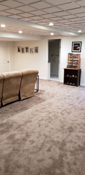 Empty beige living room with carpet, couch, wall photos, and a doorway to a lit hall