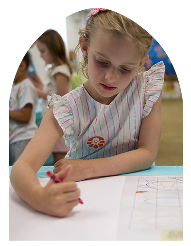 A person focused on drawing with a crayon on paper at a table, with others blurred in the background.
