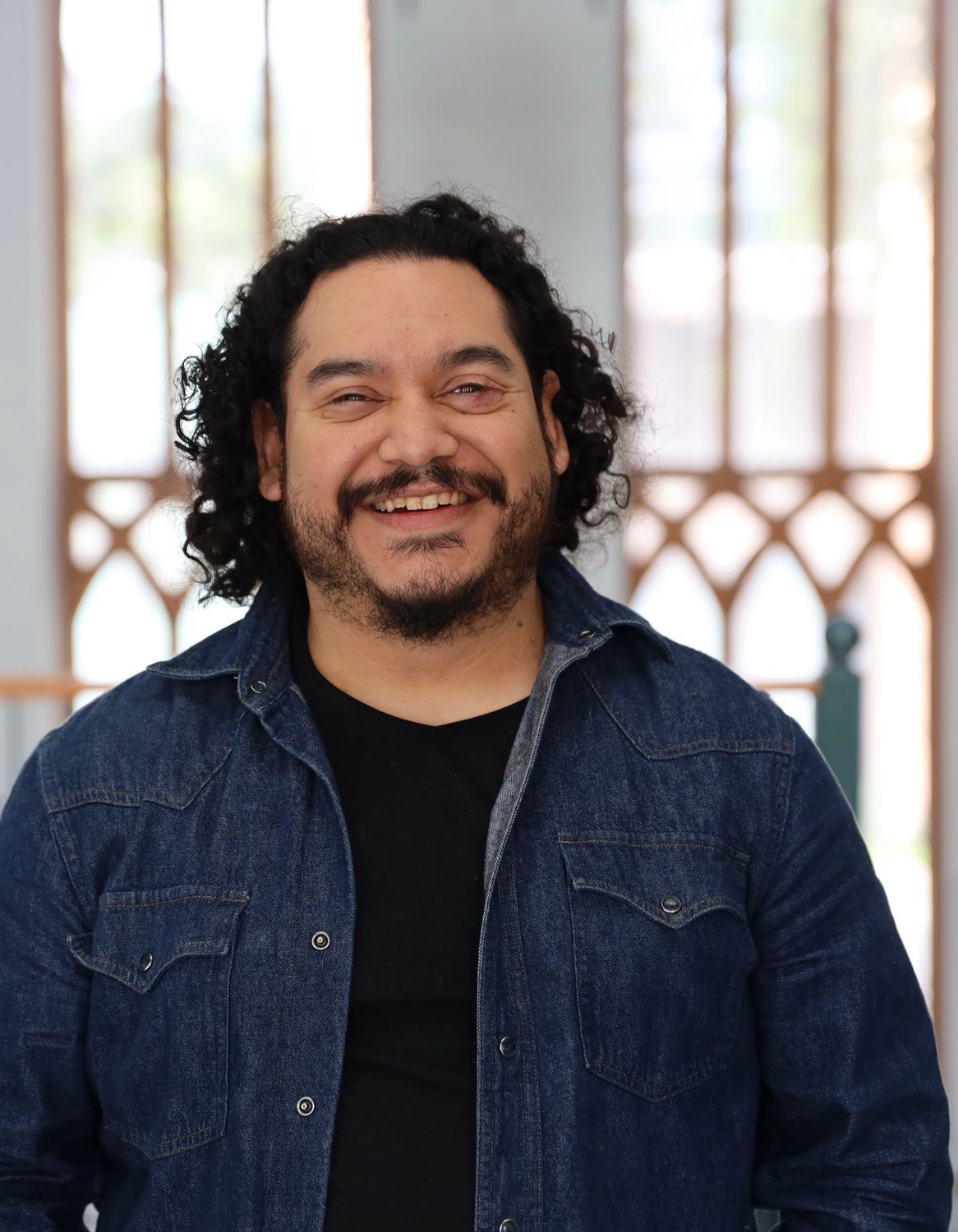 A person with dark, curly hair and a mustache smiling while wearing a dark denim button-down shirt over a black top.