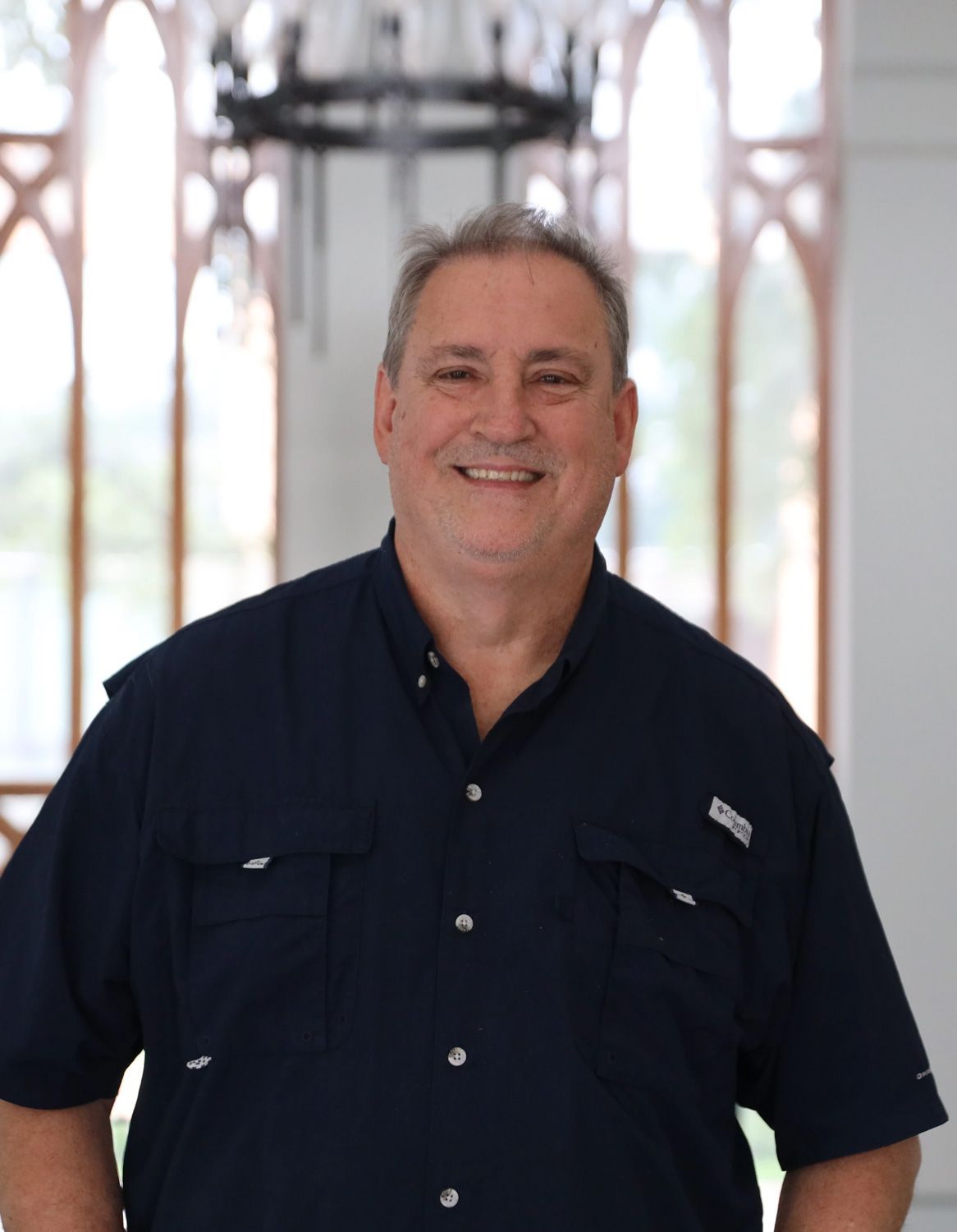 A person with short hair smiling in a dark blue short-sleeved button-down shirt in front of an arched window.