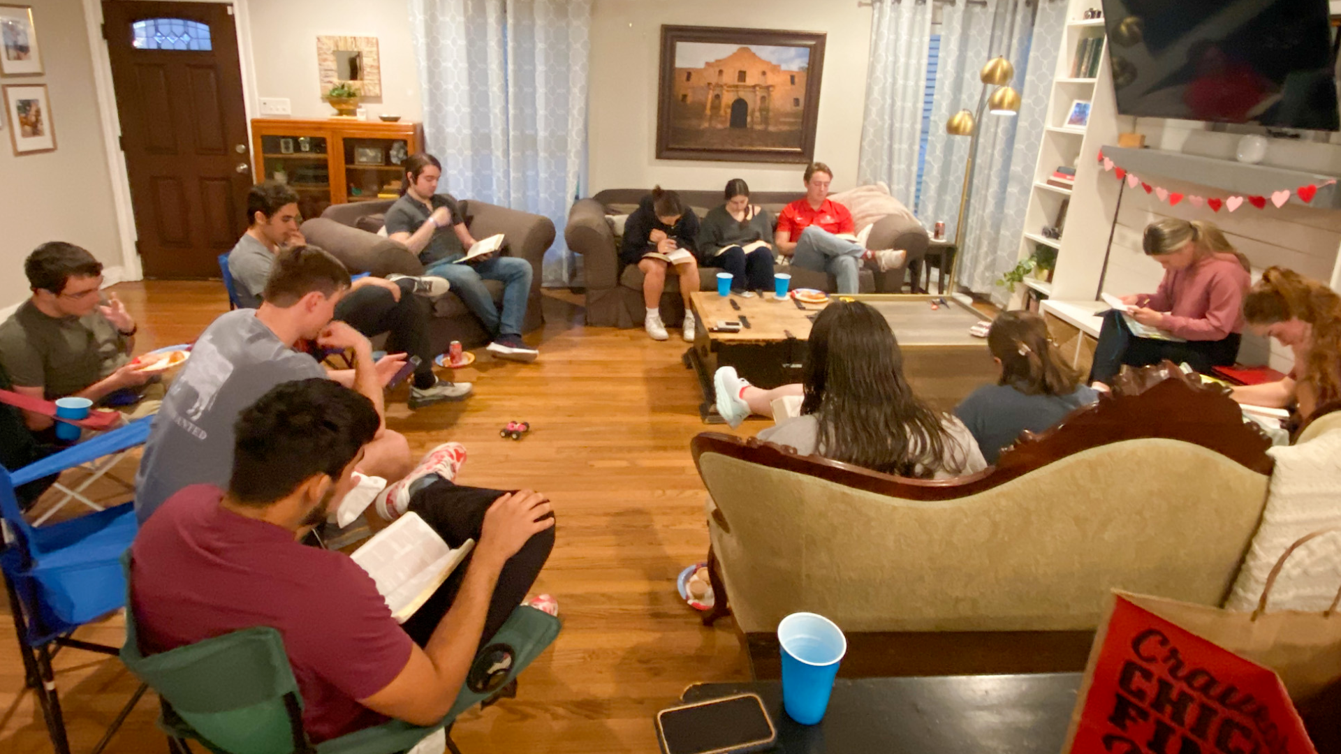 A group of people sit in a living room, some reading from books and others talking in a casual, reflective setting.