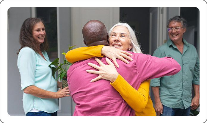 A person in a pink shirt hugs someone wearing yellow. Two other people stand by, smiling in a bright, outdoor setting.