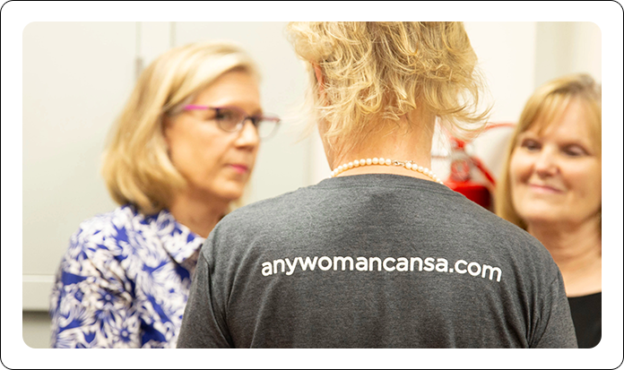 Three people interact in a room, with the person in the foreground wearing a grey shirt that reads anywomancansa.com.