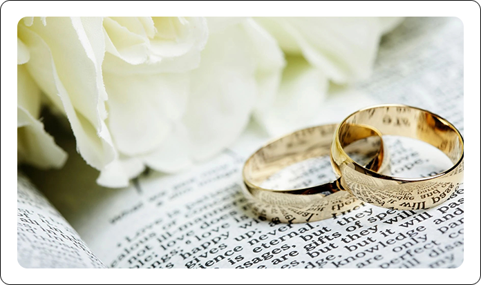 Two gold wedding rings rest on an open book next to a bouquet of white flowers.