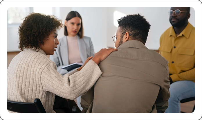 A person places a supportive hand on the shoulder of another individual in a group meeting.