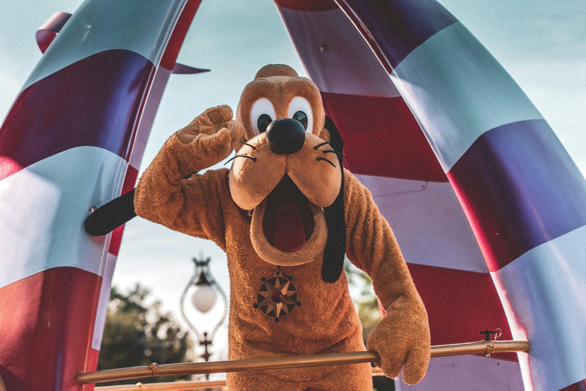 Pluto the dog character waves while standing on a colorful float at an outdoor park.