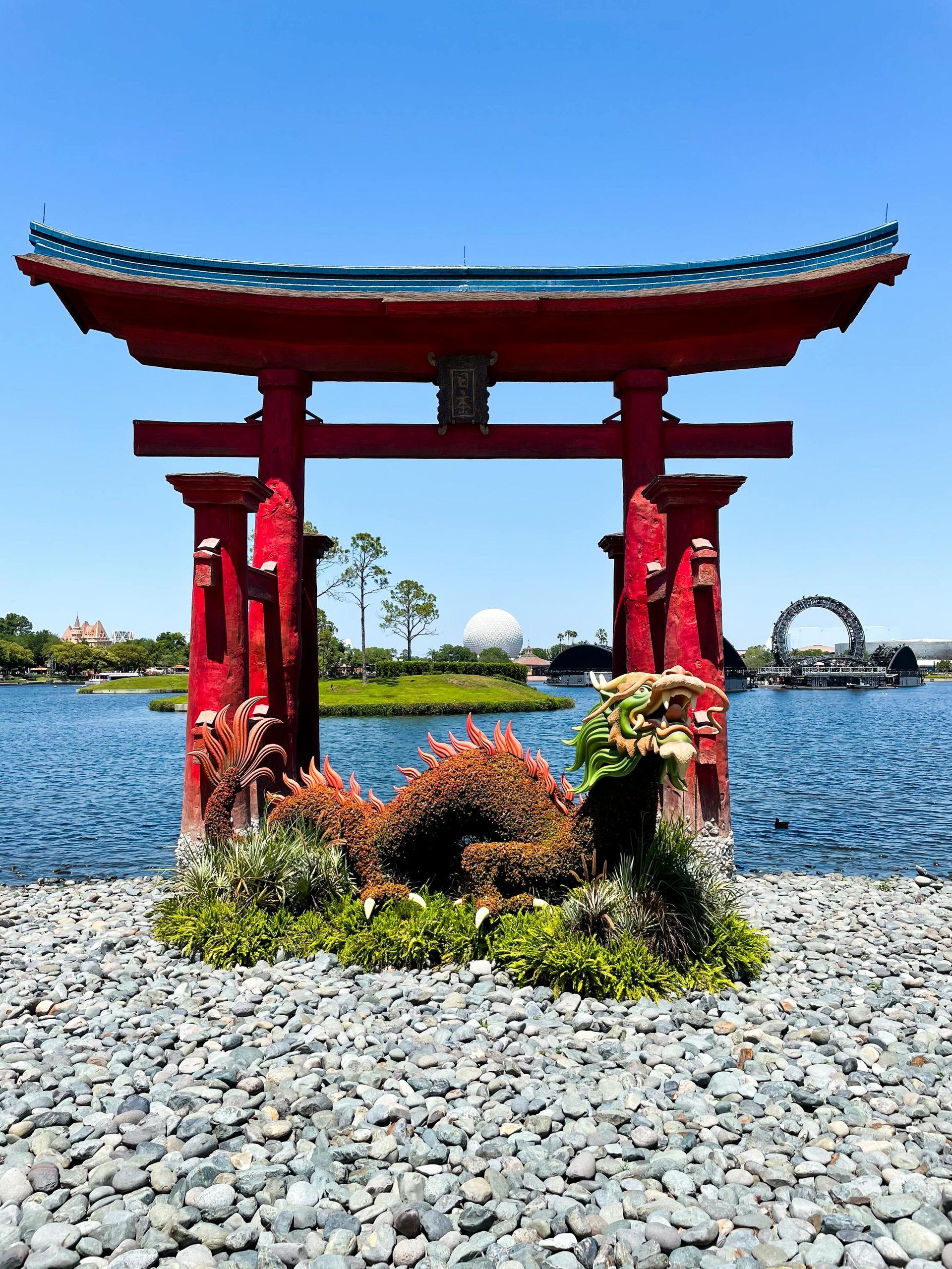 A vibrant red torii gate stands on a gravel shore overlooking a lake, with a topiary dragon and Epcot’s sphere in view.