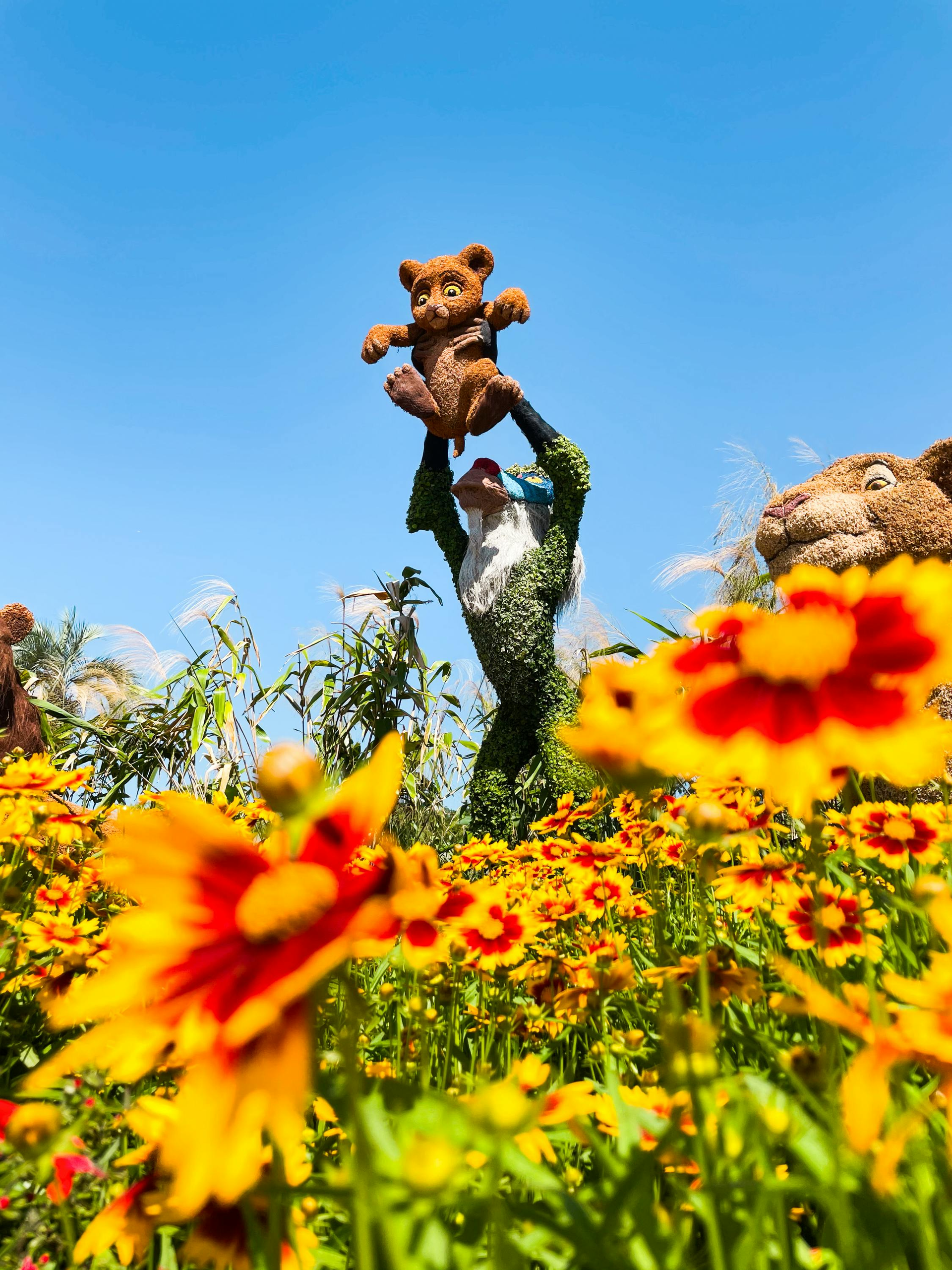 A topiary figure holds a teddy bear aloft in a vibrant field of yellow and red flowers under a clear blue sky.