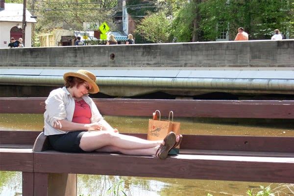 A woman is sitting on a bench near a river reading a book