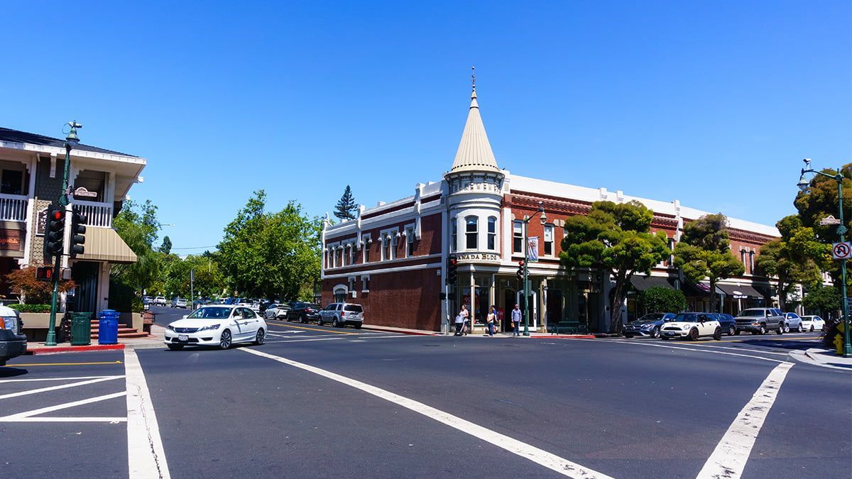 Street corner with a historic red-brick building, trees, and cars under a clear blue sky