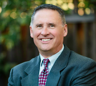 Smiling man in a suit and patterned tie outdoors with blurred greenery behind him