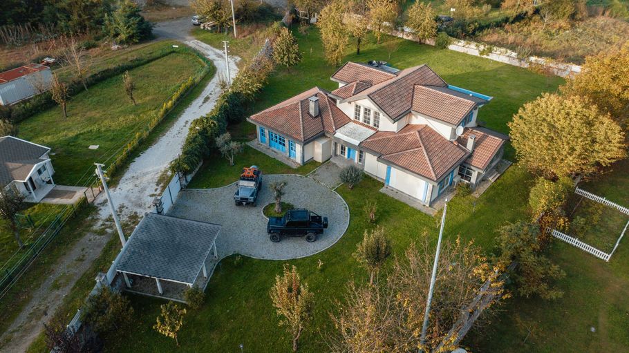 Aerial view of a house with red-tiled roofs, driveway, garage, and surrounding trees and lawns.