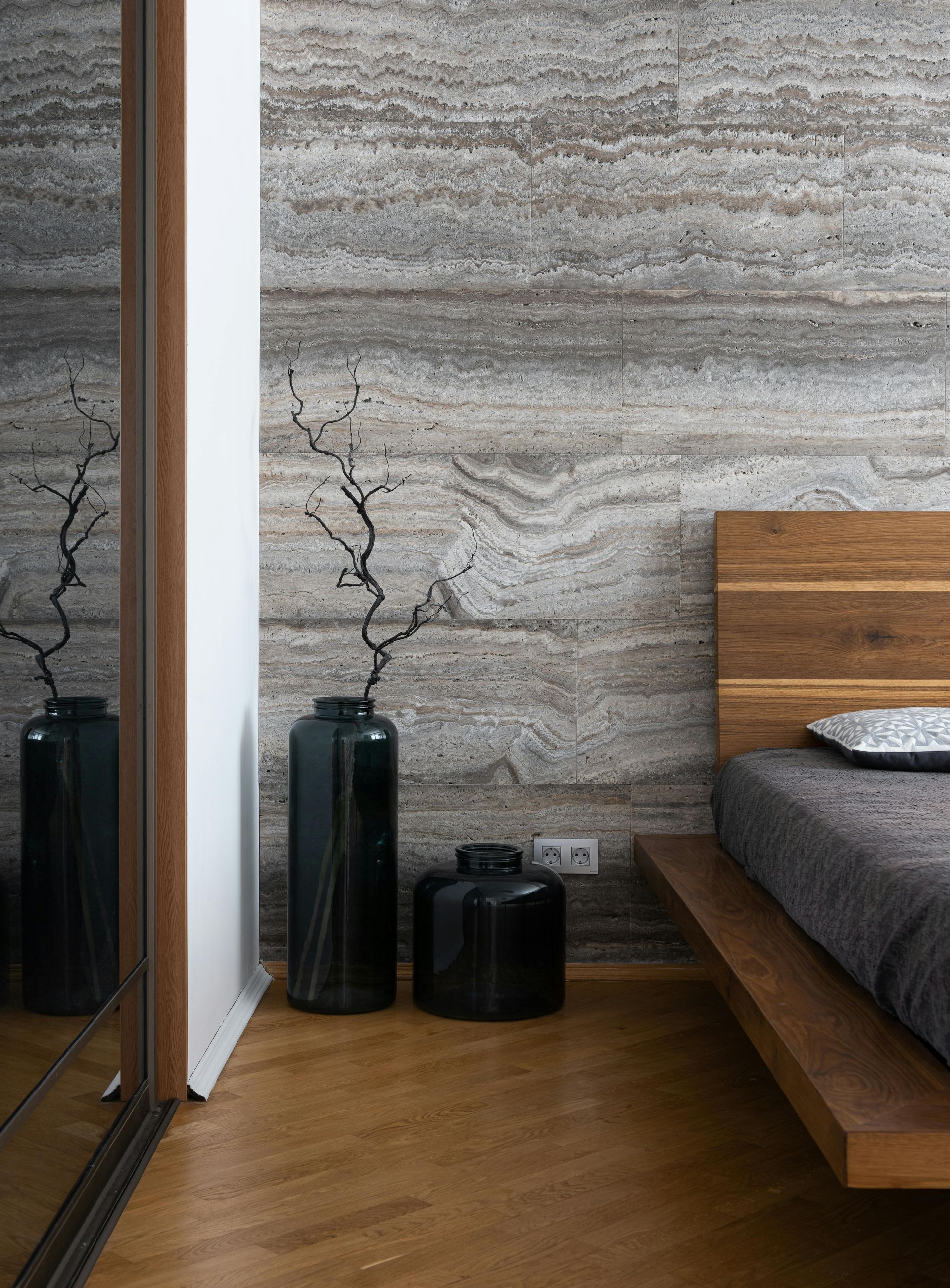 Modern bedroom with wood floor, textured stone wall, bed, mirrored closet, and tall black vases.