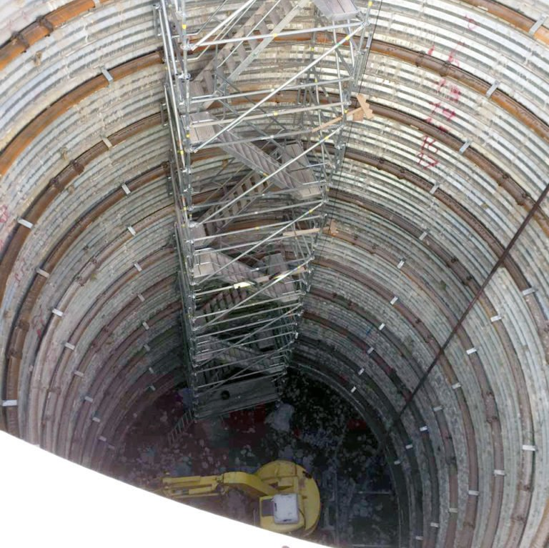 Top-down view inside a cylindrical concrete well with metal scaffolding and a yellow machine at the bottom.