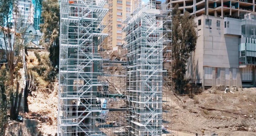 Scaffolding around a tall structure at a construction site beside rocky hills and buildings