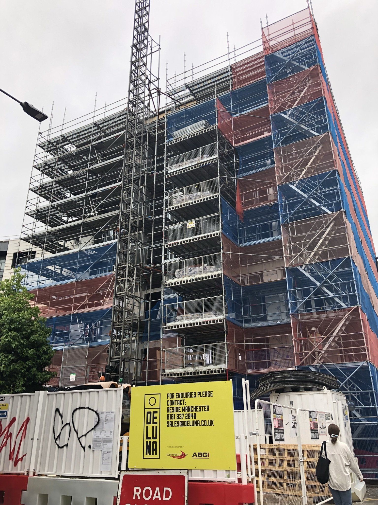 Scaffolded building under construction with blue safety netting and a crane, behind a site fence and signs