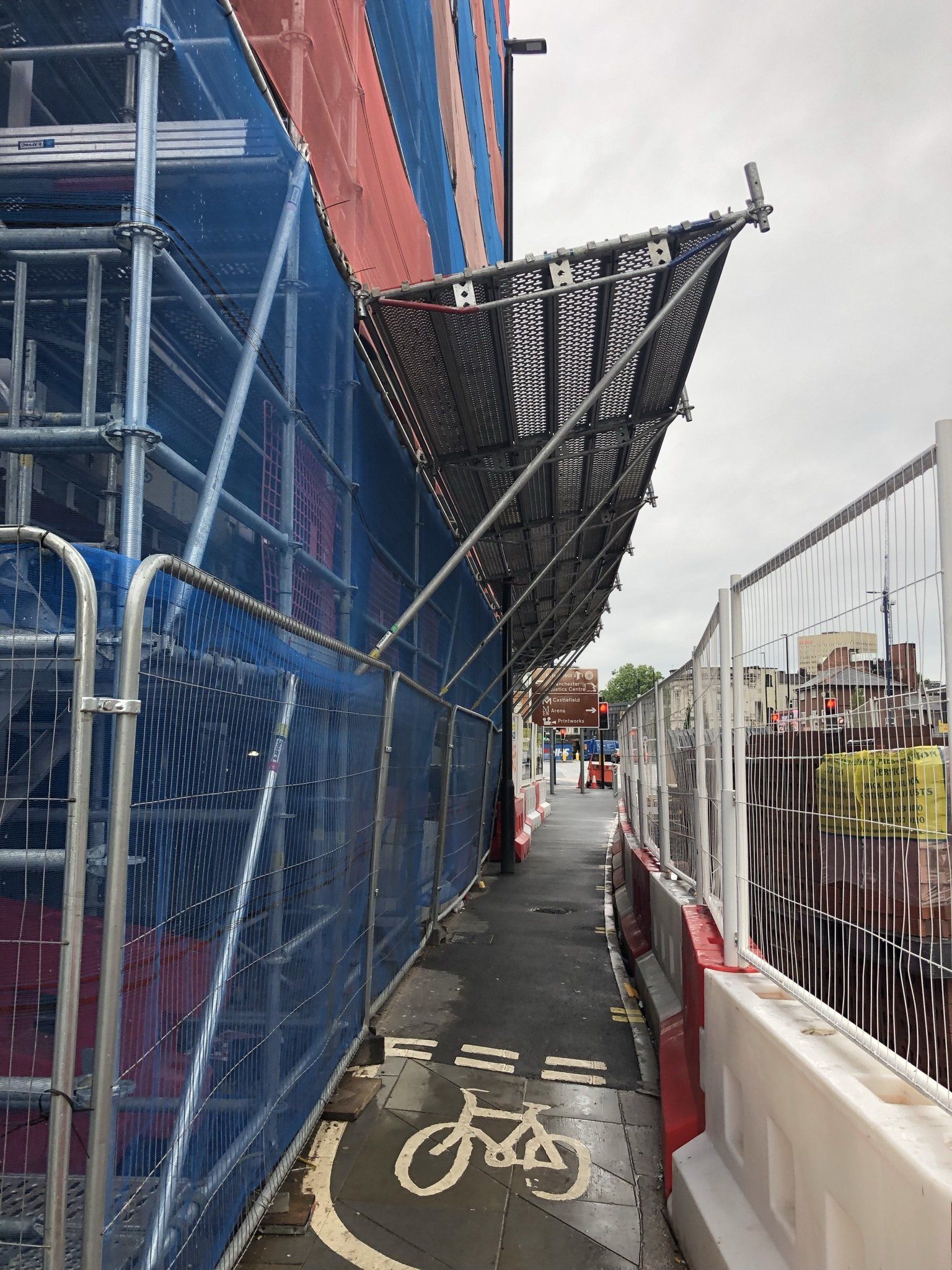 Narrow construction walkway beside blue scaffolding and fencing, with a bicycle lane symbol on the ground.