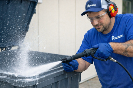 Man in blue shirt pressure-washing a black container outdoors