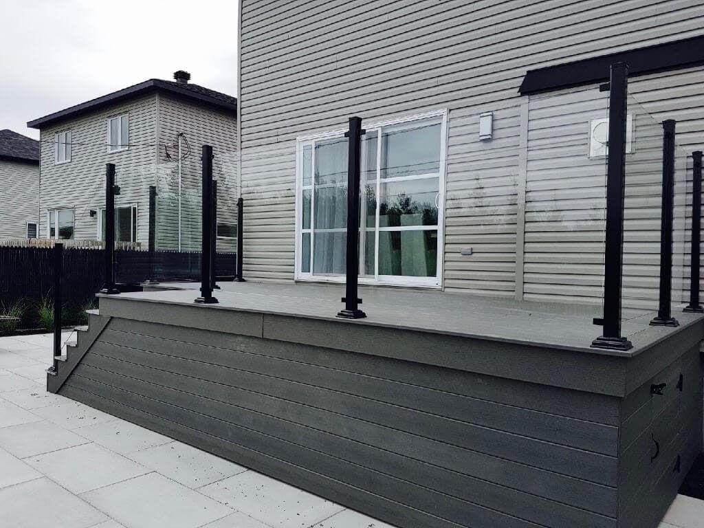 Terrasse grise surélevée avec rampes noires près de portes patio vitrées dans une cour résidentielle.