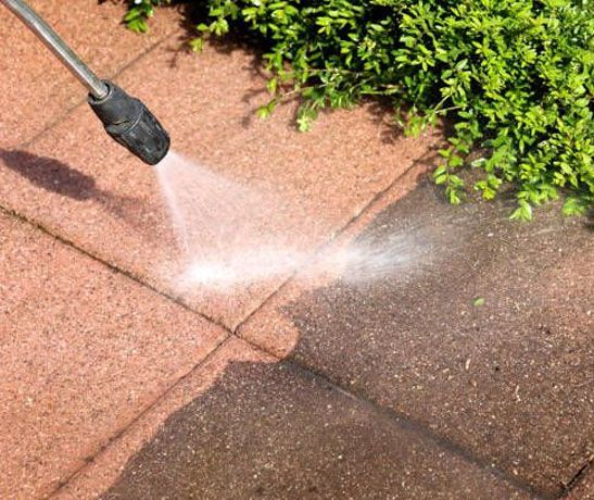 Spraying water onto a red brick patio to clean it, with green shrubs along the edge.