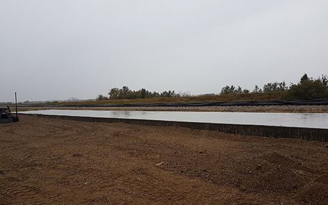Wide dirt field beside a narrow water canal under an overcast sky.