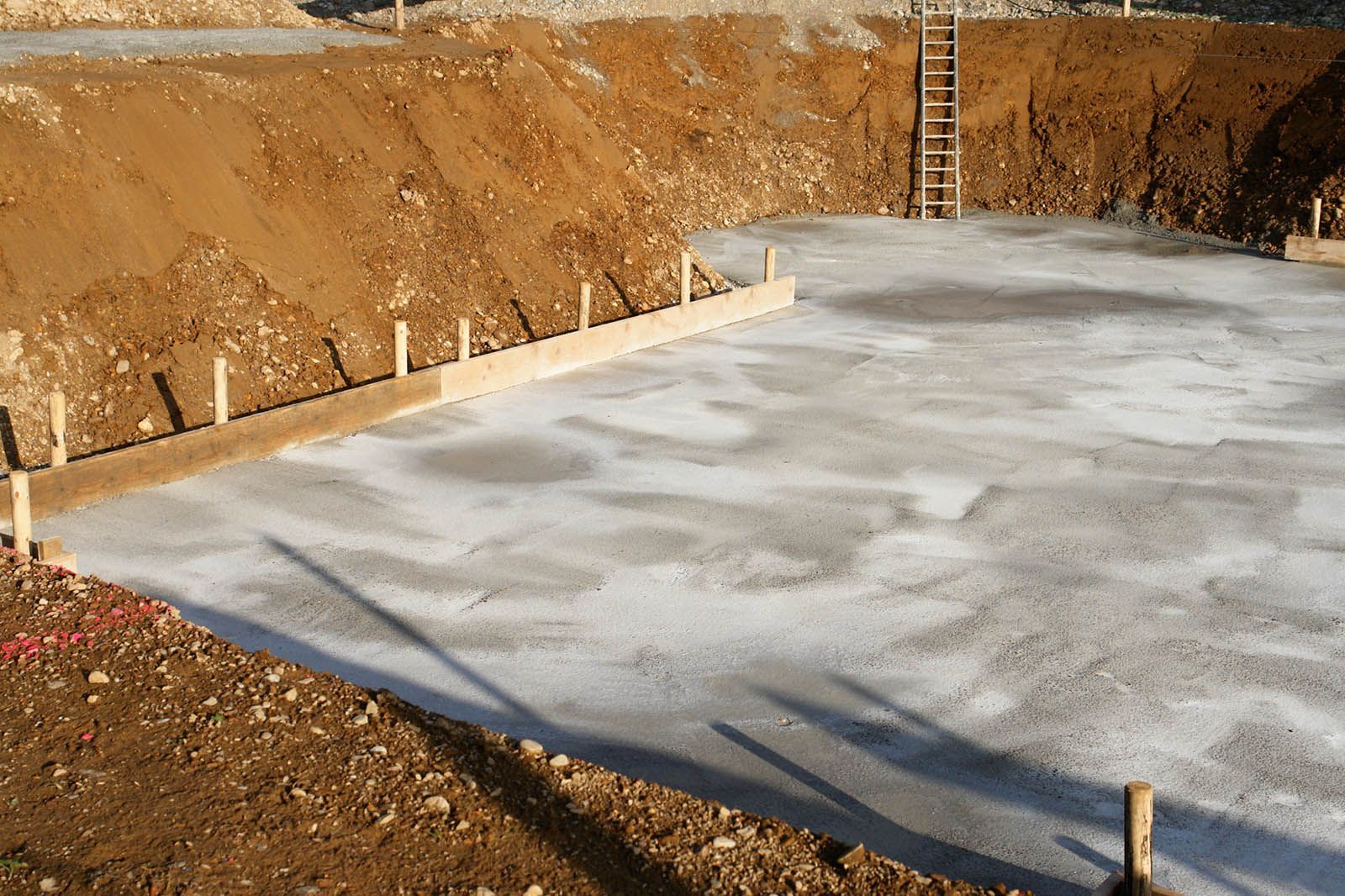 Concrete foundation slab poured in a dirt excavation site with a ladder against the back wall.