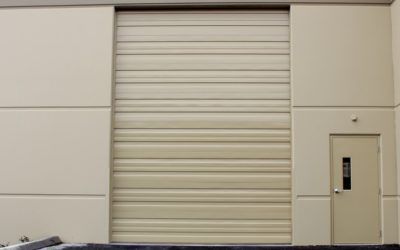 Closed beige warehouse roll-up door beside a small side door on a plain exterior wall.