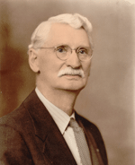 Elderly man with glasses, mustache, and suit looking forward.