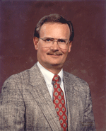 Man in a gray blazer, glasses, and tie smiling in front of a maroon background.