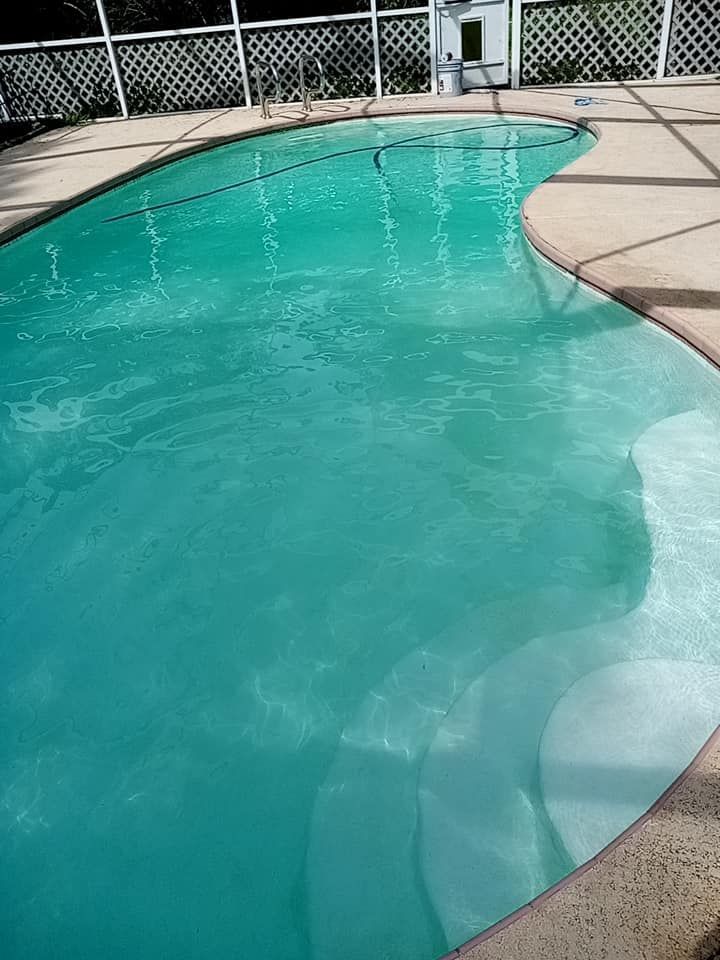 A turquoise swimming pool with steps, surrounded by a light-colored deck and fence.