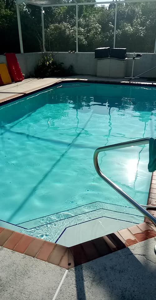 Swimming pool with brick border, steps, and handrail, surrounded by a screened enclosure and trees.