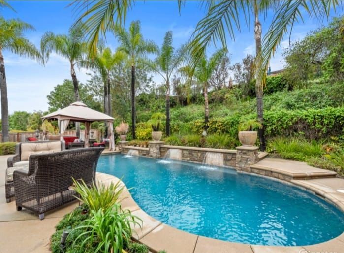 Backyard pool with waterfalls, gazebo, and lounge seating; lush greenery, palm trees, blue water.