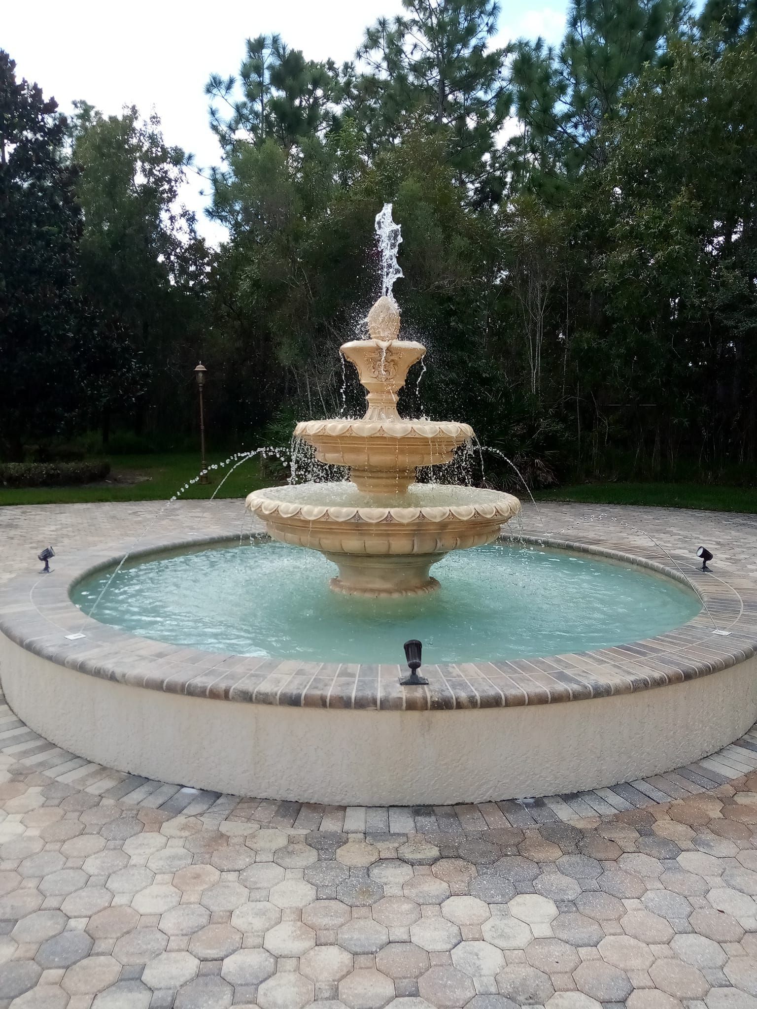 Tiered stone fountain with water spraying in a round pool, surrounded by brick pavers, trees in the background.
