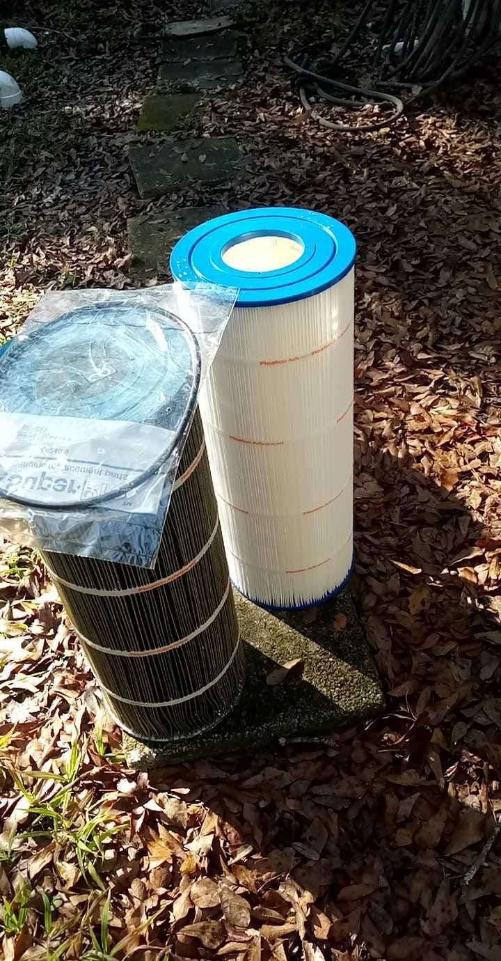Two cylindrical pool filters on the ground; one black, one white with a blue top, among leaves and branches.