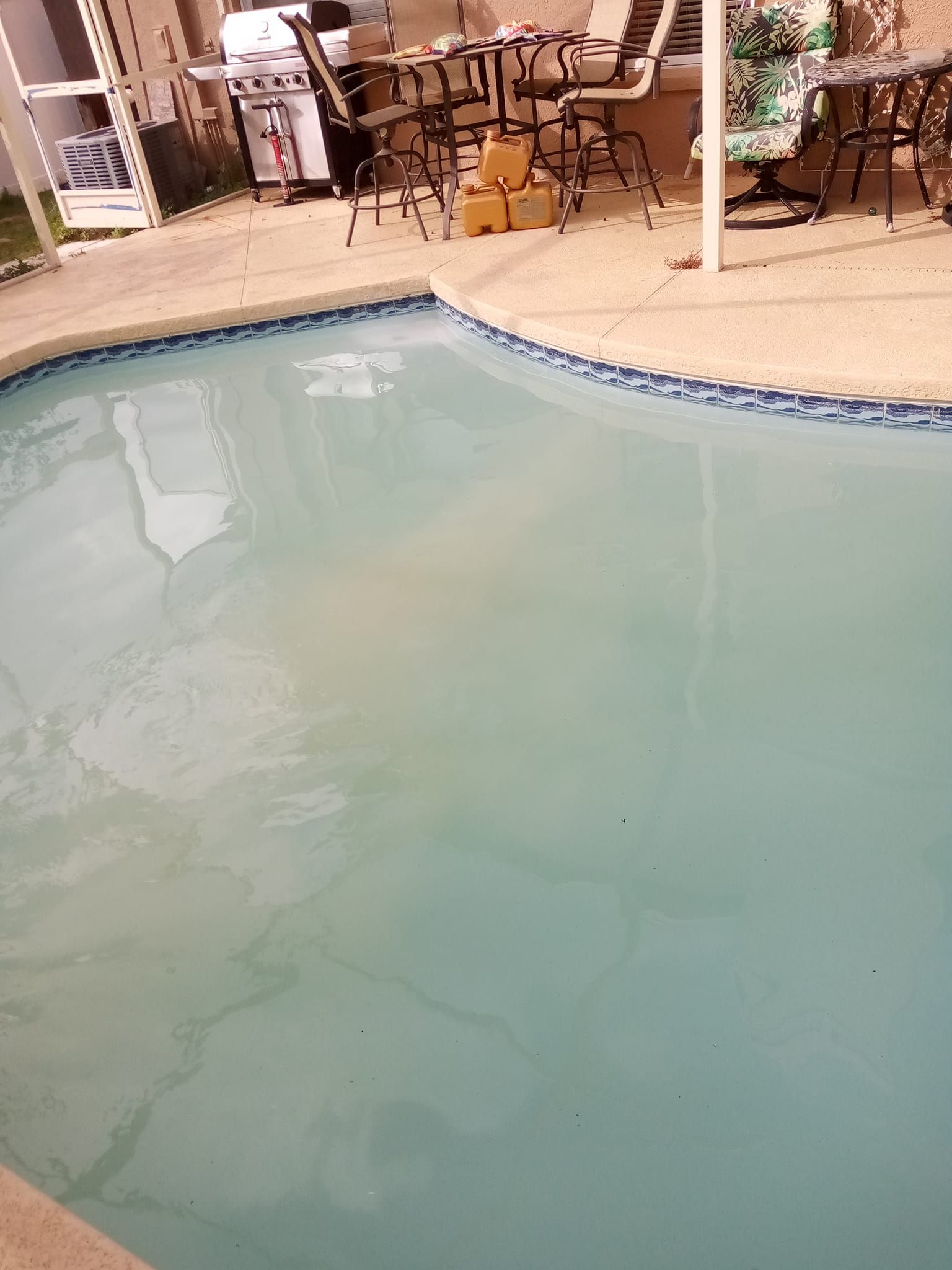 Pool filled with cloudy blue water, surrounded by concrete deck. Outdoor setting with grill and chairs.
