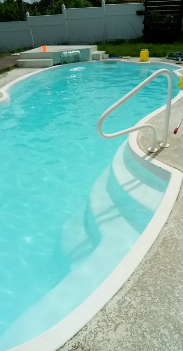 Swimming pool with white steps and handrail, surrounded by concrete and a white fence.
